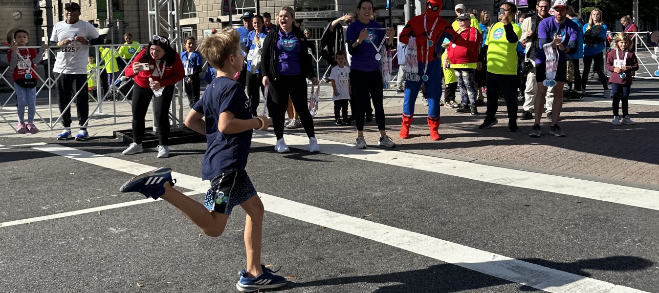 Child running toward the finish line being cheered on by spectators, including one dressed as Spider Man holding medals