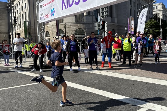 Child running toward the finish line being cheered on by spectators, including one dressed as Spider Man holding medals